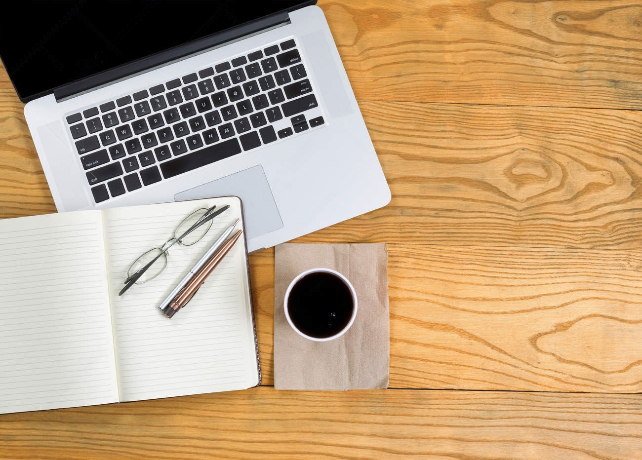 Ordenador sobre una mesa con cuaderno, bolígrafo, gafas y taza de café